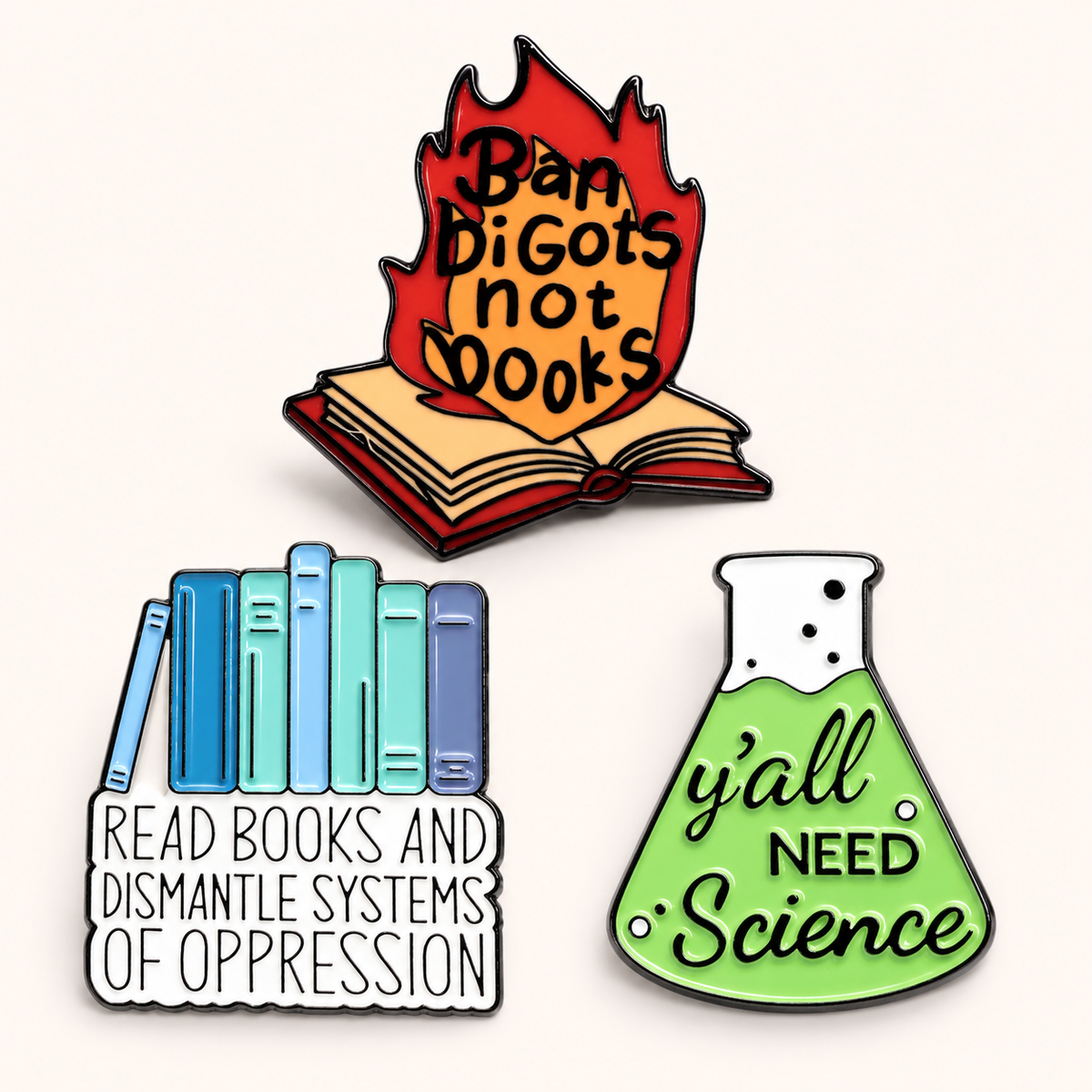 Three enamel pins displayed on a light cream background: a top pin shows an open book engulfed in flames with the text 'Ban DiGots not Books'; a bottom-left pin shows blue and teal books with the text 'READ BOOKS AND DISMANTLE SYSTEMS OF OPPRESSION'; a bottom-right pin shows a green beaker with the text 'y'all NEED Science'.