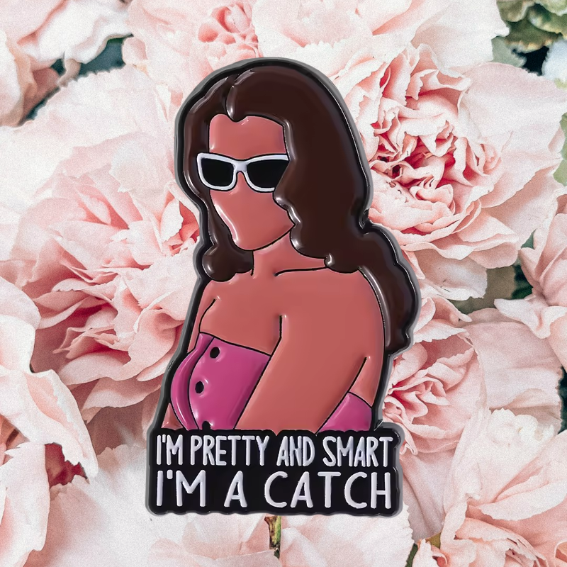 An enamel pin shaped like a woman with brown hair, white sunglasses, and a pink top, with the text 'I'M PRETTY AND SMART I'M A CATCH' at the bottom, displayed on a background of pink flowers.