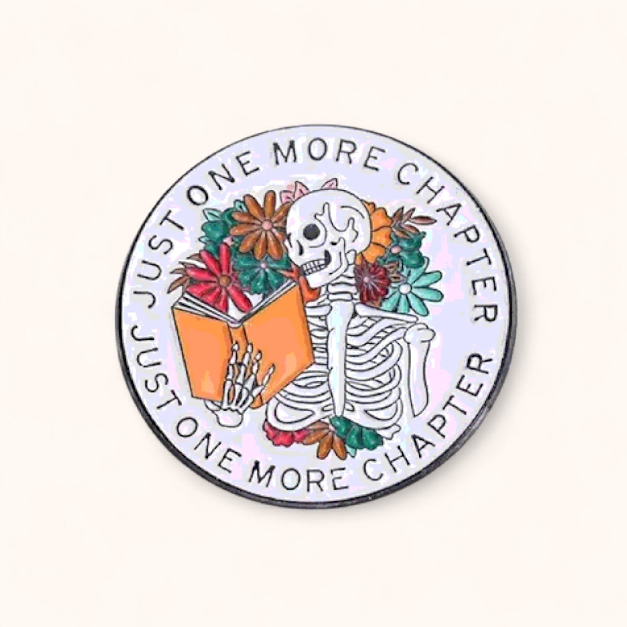 Circular enamel pin with skeleton holding a book and floral elements, text 'Just One More Chapter'.