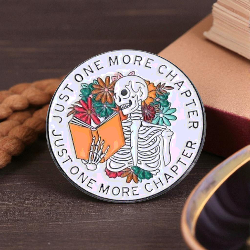 Circular badge with skeleton and book design, text 'Just One More Chapter' on a wooden surface.