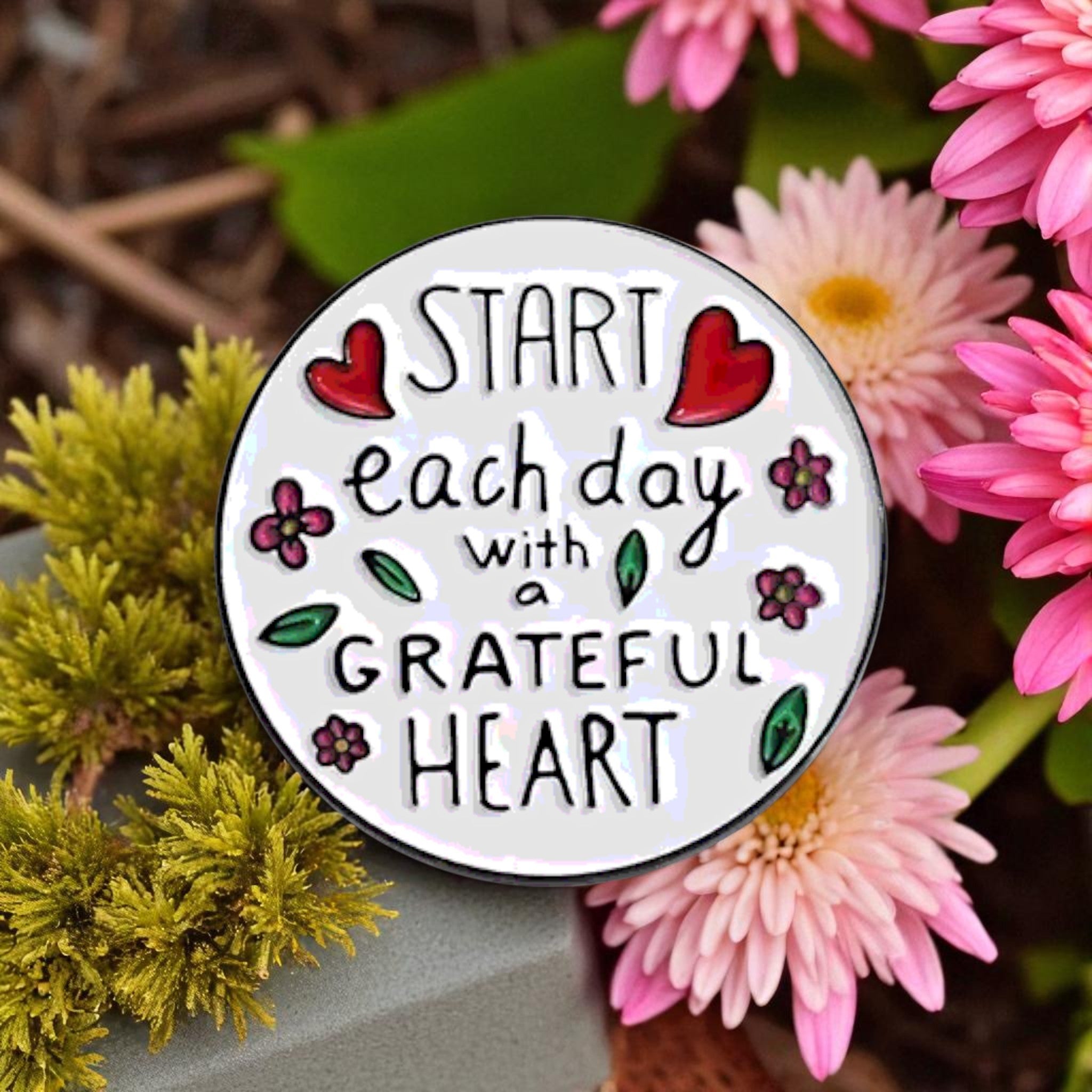 Circular enamel pin with 'Start each day with a grateful heart' text surrounded by pink flowers.