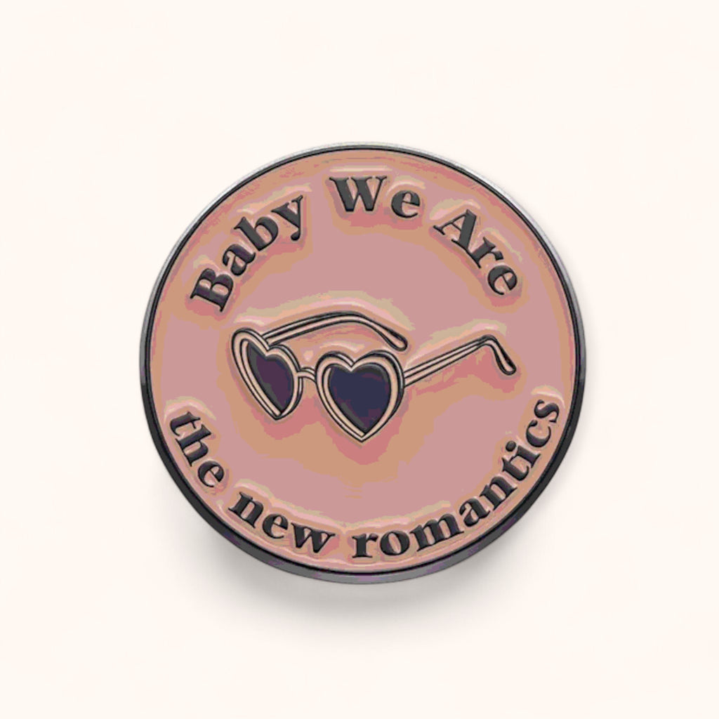 Round enamel pin with sunglasses design and text 'Baby We Are the new romantics' on a pink background
