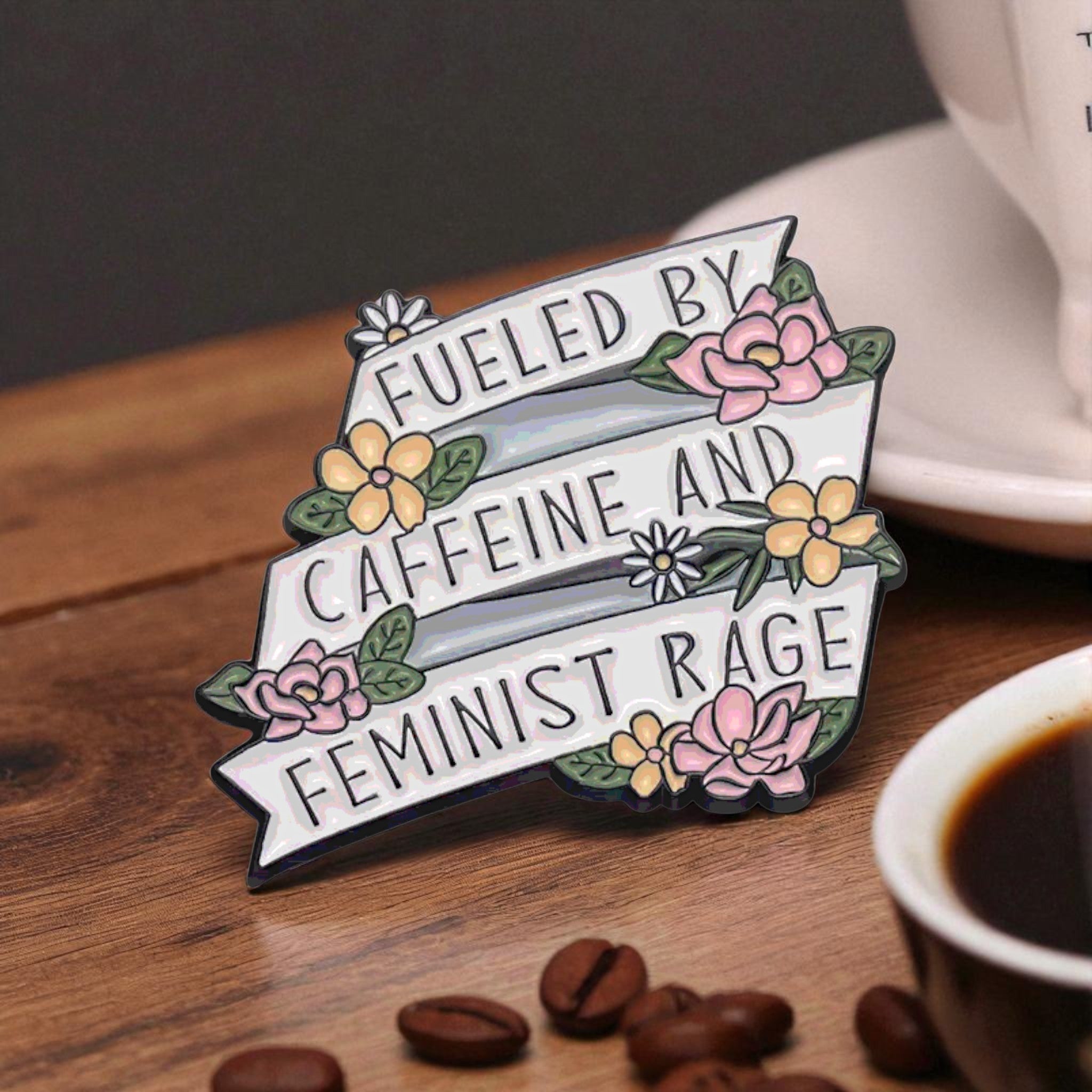 Enamel pin with 'Fueled by caffeine and feminist rage' text on a wooden surface with coffee beans and a cup of coffee.